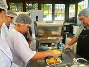 mulher com guarda-pó e touca branco em cozinha industrial, prato com comida, professor em frente auxiliando.