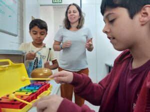 A imagem mostra três pessoas. À direita um adolescente moreno toca uma miniatura em brinquedo de um piano amarelo. O garoto é moreno e usa moleton marrom. Ao centro uma mulher branca, cabelos loiros.