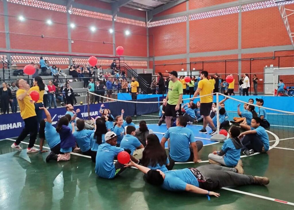 Imagem de crianças sentadas em ginásio com rede de vôlei baixa e texto - Festival Paralímpico terá 2ª edição em Florianópolis no próximo sábado, 07